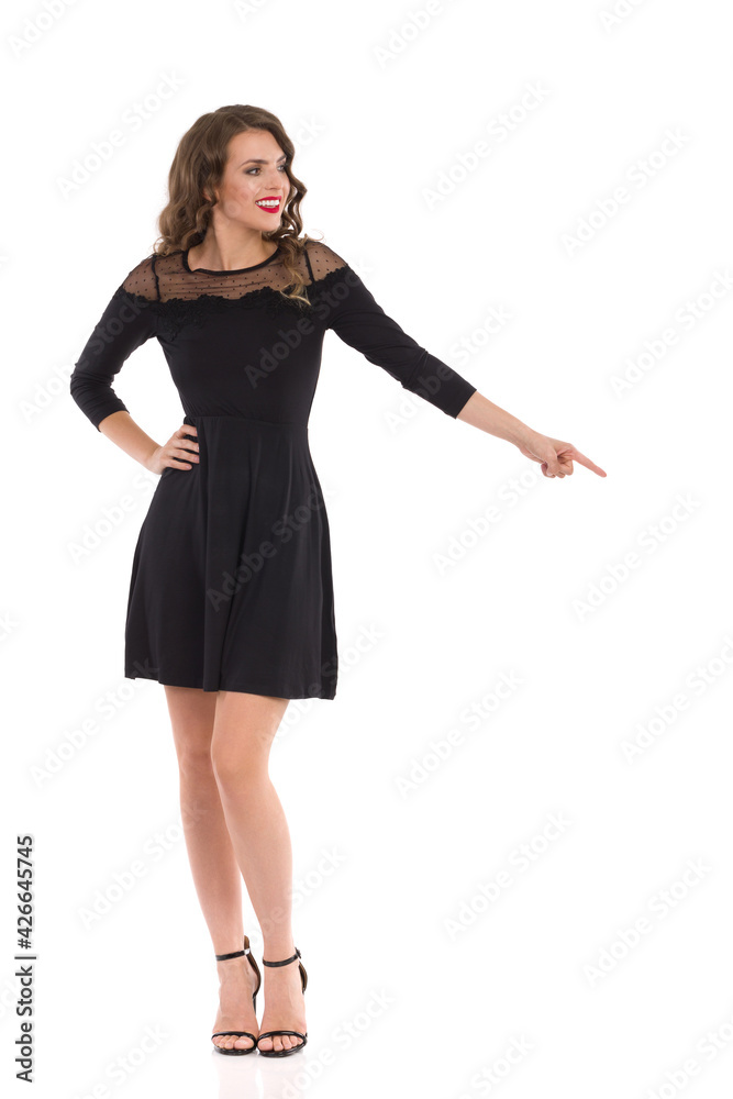 Elegant Young Woman In Black Cocktail Dress And High Heels Is Standing And Pointing Down Elegant Young Woman In Black Cocktail Dress And High Heels Is Standing And Pointing Down