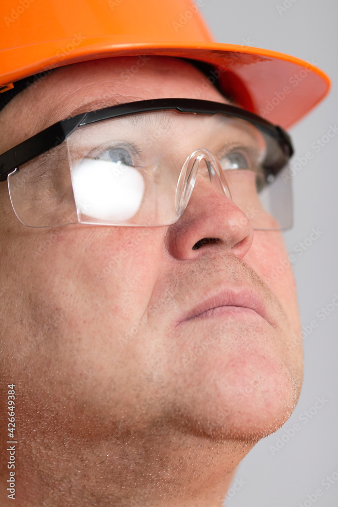 face of confident plump engineer, middle-aged carchitect in hardhat at and protective glasses looking up on grey studio background face of confident plump engineer, middle-aged carchitect in hardhat at and protective glasses looking up on grey studio background