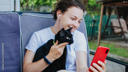Cheerful young woman wearing headphones holds black pet cat using smartphone for video call, gesturing hi to friend or parent, sitting on sofa at home backyard outdoors. Sharing data on social media