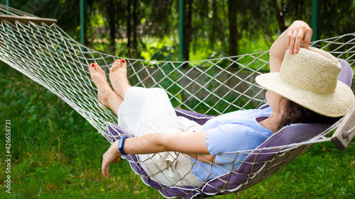 Don't disturb me. Beautiful young woman relaxing in hammock, covering face with summer straw hat. Enjoying hot sunny day in shadow, resting lying in hummock in green garden outdoors. Weekend concept