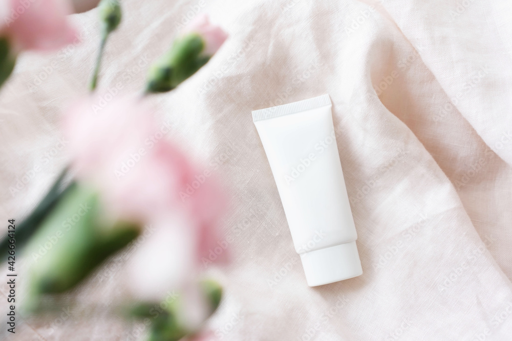 white cosmetic tube with face cream, body lotion or cleanser  on a light fabric background with pink flowers. Sensitive skin care concept. Copy space, mock up