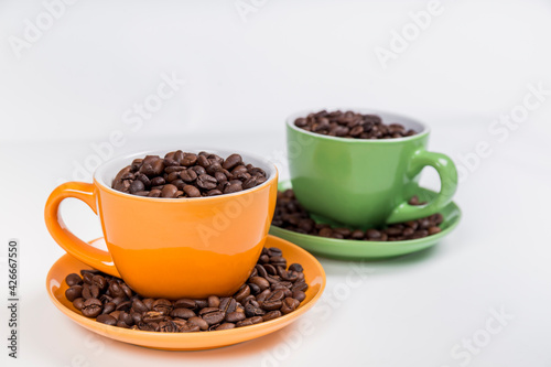 set of two ceramic cups with their plates one orange and one green filled with coffee beans  on white background  horizontal photo