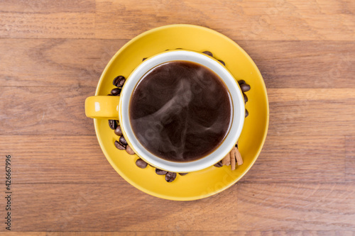 coffee composition, aerial shot of a yellow ceramic cup full of single coffee, on the coffee plate there are coffee beans and a cinnamon stick, the background surface is wooden, horizontal photo