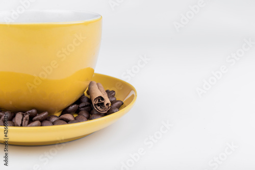 coffee composition, close-up of part of a yellow ceramic cup with its plate full of coffee beans and a piece of cinnamon stick on a white background, horizontal photo with space to write