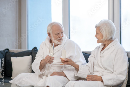 Senior couple in white robes drinking champagne and looking pleased © zinkevych