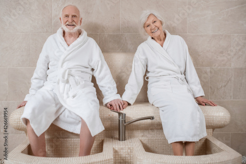 Senior couple in white robes having procedures for legs in spa salon