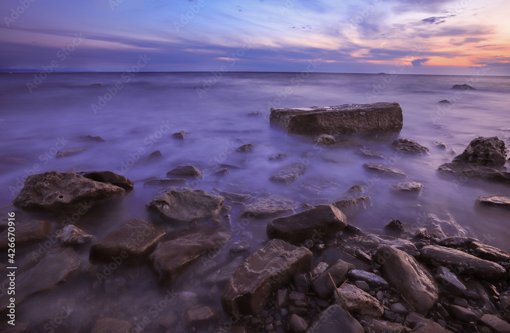 Rocky seascape at sunrise with waves flowing over harsh granite rocks. Sunset over dark rocks at the coast. A long exposure image with waves flowing over rocky outcrops. Violet sea. Mystic scene.