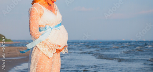 Pregnant woman with hands on her stomach, in the summer by the sea in a white dress