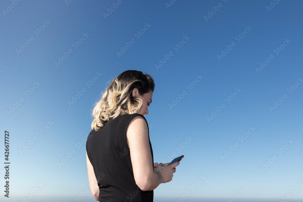 young blonde woman walking on the beach looking at the phone on a sunny spring day with blue sky