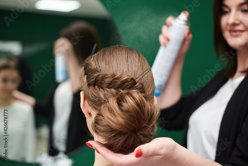 Bride getting ready for wedding. Professional hairdresser making coiffure for female client, applying hairspray in front of big mirror. Work process in beauty studio. Close-up picture of gala hairdo.