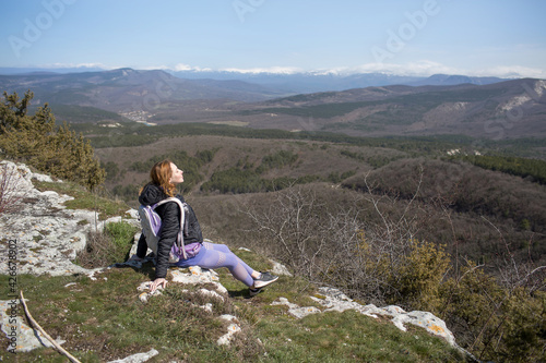 A girl in sports clothes sits on the top of a mountain with her back to the camera and looks into the distance at a beautiful landscape