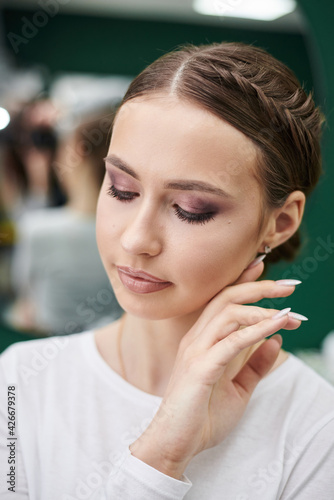 Young girl with fancy hairdo and make-up posing in front of mirror in beauty studio. Close-up portrait of makeup fashion model. Love yourself female concept. Bride's morning on wedding day.