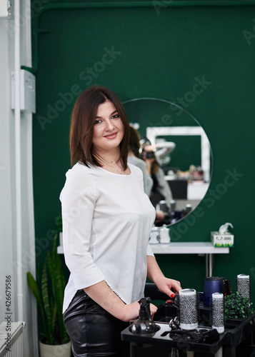 Portrait of professional hairdresser in beauty salon. Young brunette female hair stylist standing at her workplace with accessories near big mirror. Work process in barber shop.