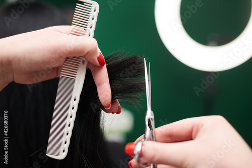 Professional hairdresser making haircut for female client. Close-up picture of hair stylist's hands holding scissors and comb, cutting shiny black hair. Work process in barber shop.