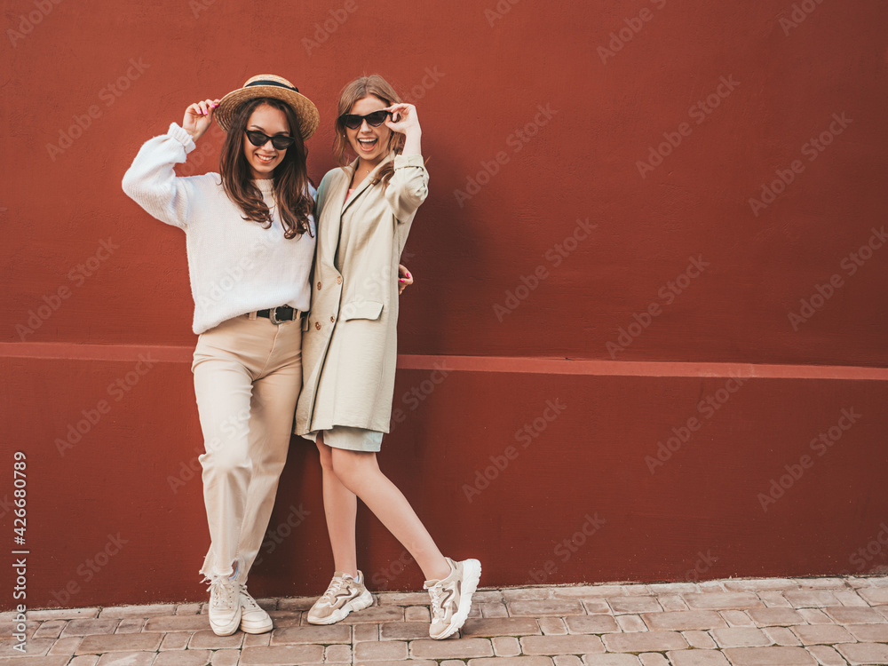 Two young beautiful smiling hipster female in trendy white sweater and coat. Sexy carefree women posing near red wall in the street in hat. Positive models having fun in sunglasses