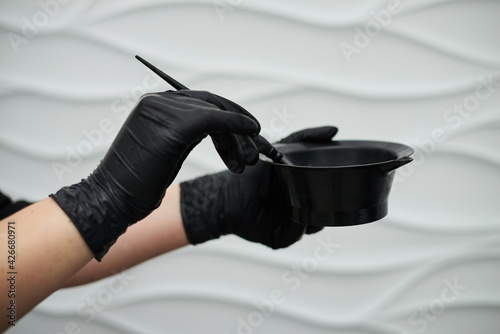 Close-up picture of hairdresser's tools in female hands with black gloves. Process of mixing hair dye with brush in plastic bowl. Hair stylist preparing for dying hair in barber shop. Beautification.