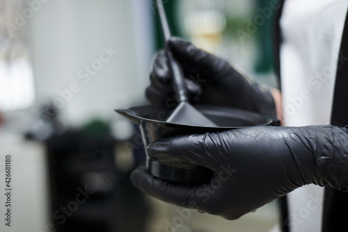 Close-up picture of hairdresser's tools in female hands with black gloves. Process of mixing hair dye with brush in plastic bowl. Hair stylist preparing for dying hair in barber shop. Beautification.