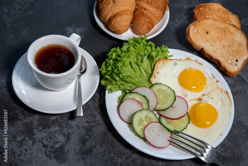 Two fried eggs, a leaf of lettuce, slices of cucumber and radish on a white plate, toast, croissants and a cup of tea or coffee. Breakfast. Healthy eating. Close-up