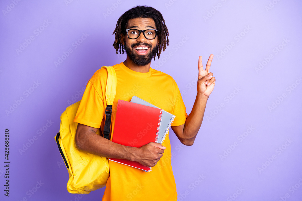 Portrait of nice impressed brunet guy hold books show v-sign wear spectacles bag orange t-shirt isolated on vibrant lilac background Portrait of nice impressed brunet guy hold books show v-sign wear spectacles bag orange t-shirt isolated on vibrant lilac background