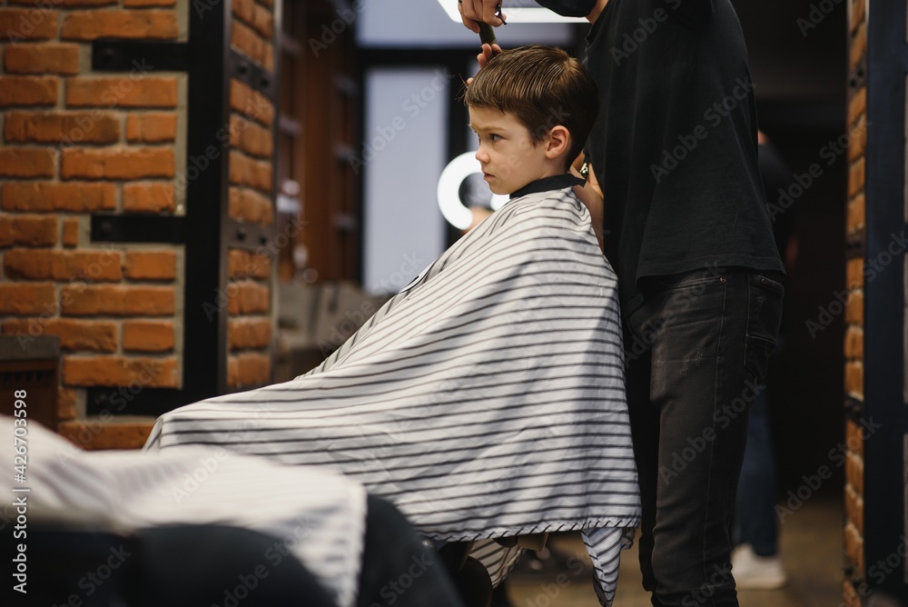 Children hairdresser with scissors is cutting little boy against a dark background. Contented cute preschooler boy getting the haircut.