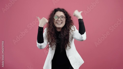 Happy young woman with curly hair, with her hands shows her thumb up as a sign, recommending smiling. Young hipster in white jacket and black shirt, with glasses posing isolated on pink background in photo