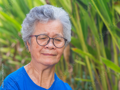Portrait of a senior woman with short gray hair wearing glasses and looking down while standing in a garden. Space for text. Concept of aged people and healthcare
