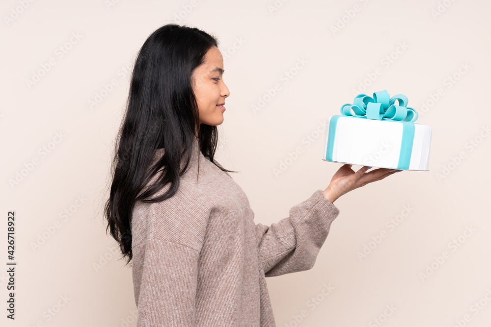 Teenager Asian girl holding a big cake isolated on beige background with happy expression Teenager Asian girl holding a big cake isolated on beige background with happy expression