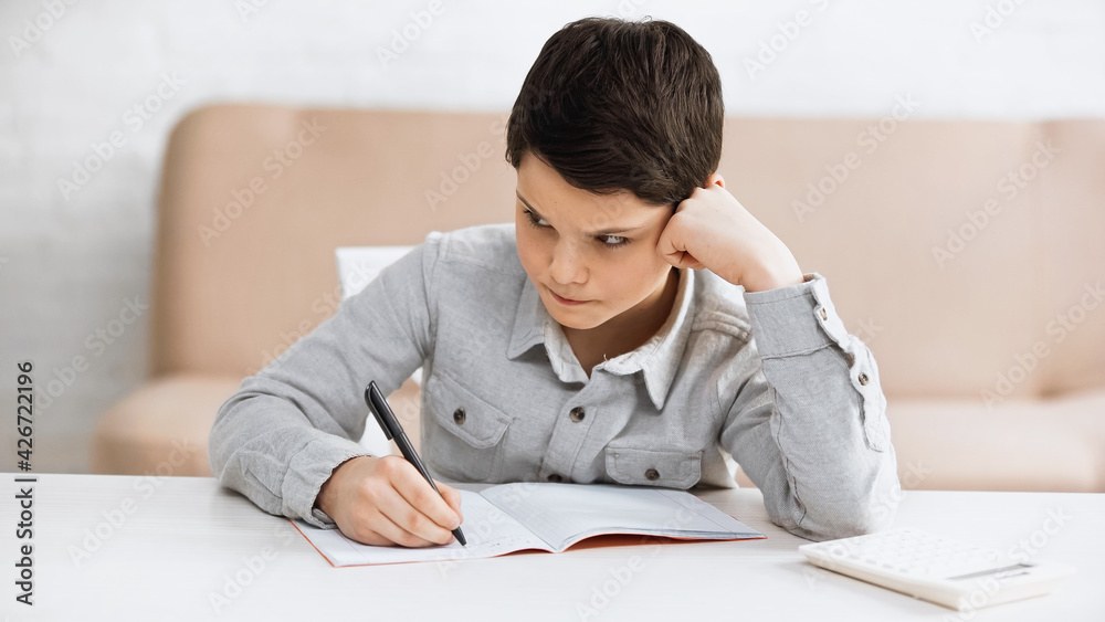 Pensive boy writing on notebook near calculator on blurred foreground at home. Pensive boy writing on notebook near calculator on blurred foreground at home.