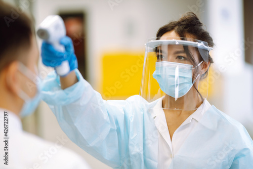 Young nurse in visor and protective gloves using infrared digital thermometer check temperature measurement on the forehead during the coronavirus pandemic. 