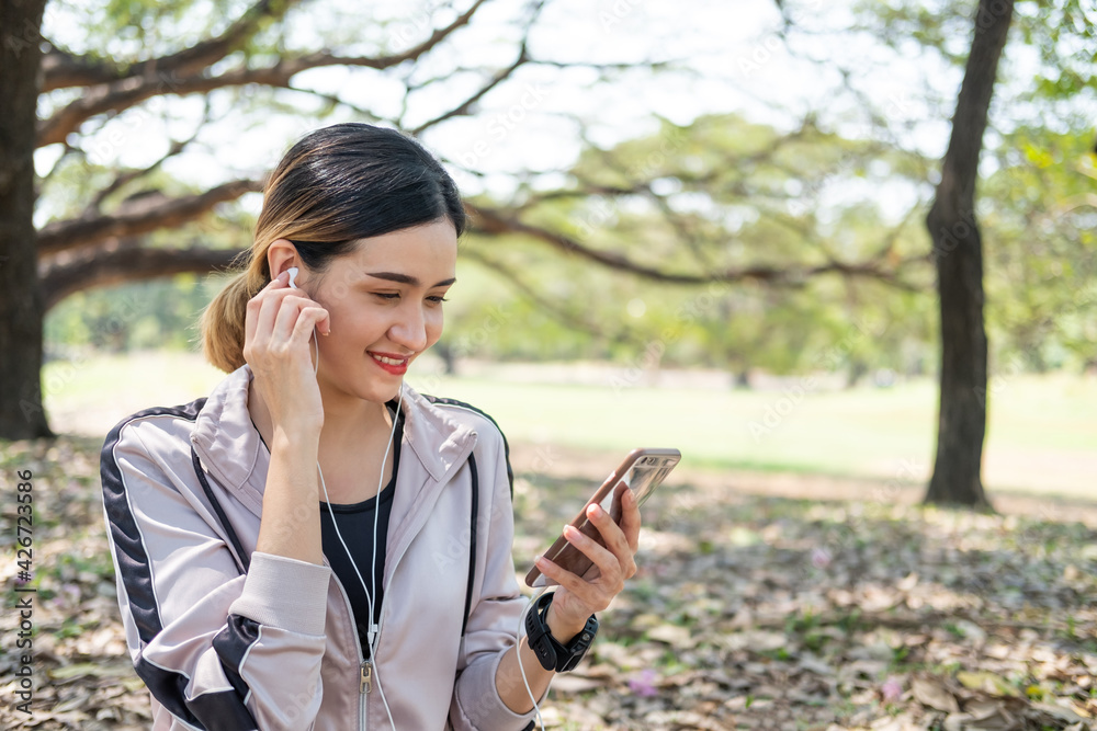 Selective focus at face of young beautiful Asian women using smart watch to track activity and listen music from smartphone while warm up before exercise yoga with natural and trees at background. Selective focus at face of young beautiful Asian women using smart watch to track activity and listen music from smartphone while warm up before exercise yoga with natural and trees at background.
