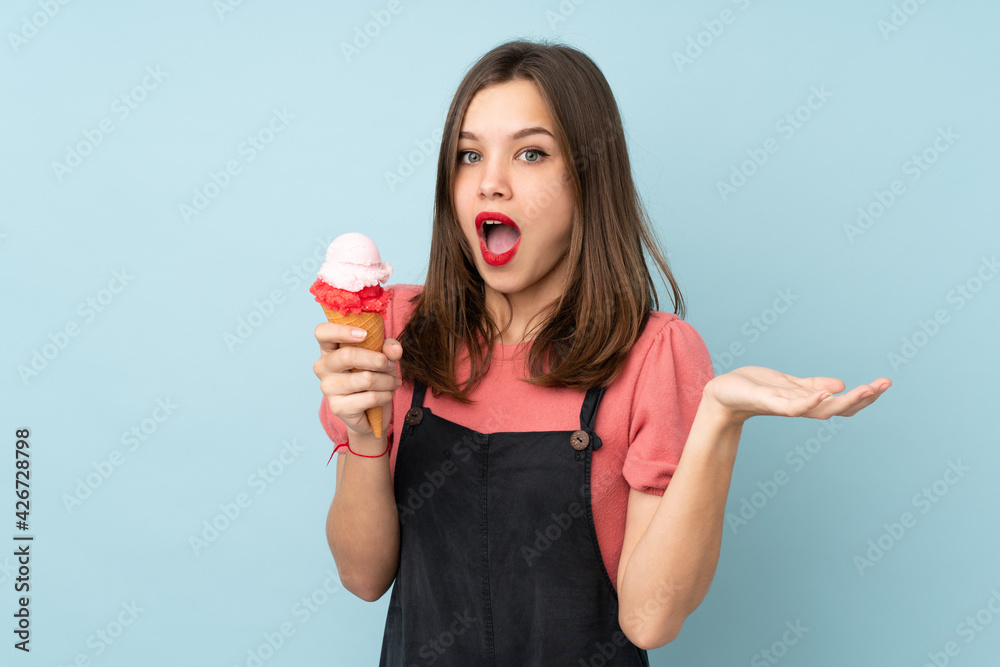 Teenager girl holding a cornet ice cream isolated on blue background with shocked facial expression