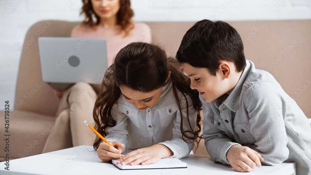 Kids doing homework on notebook near mother with laptop on blurred background. Kids doing homework on notebook near mother with laptop on blurred background.