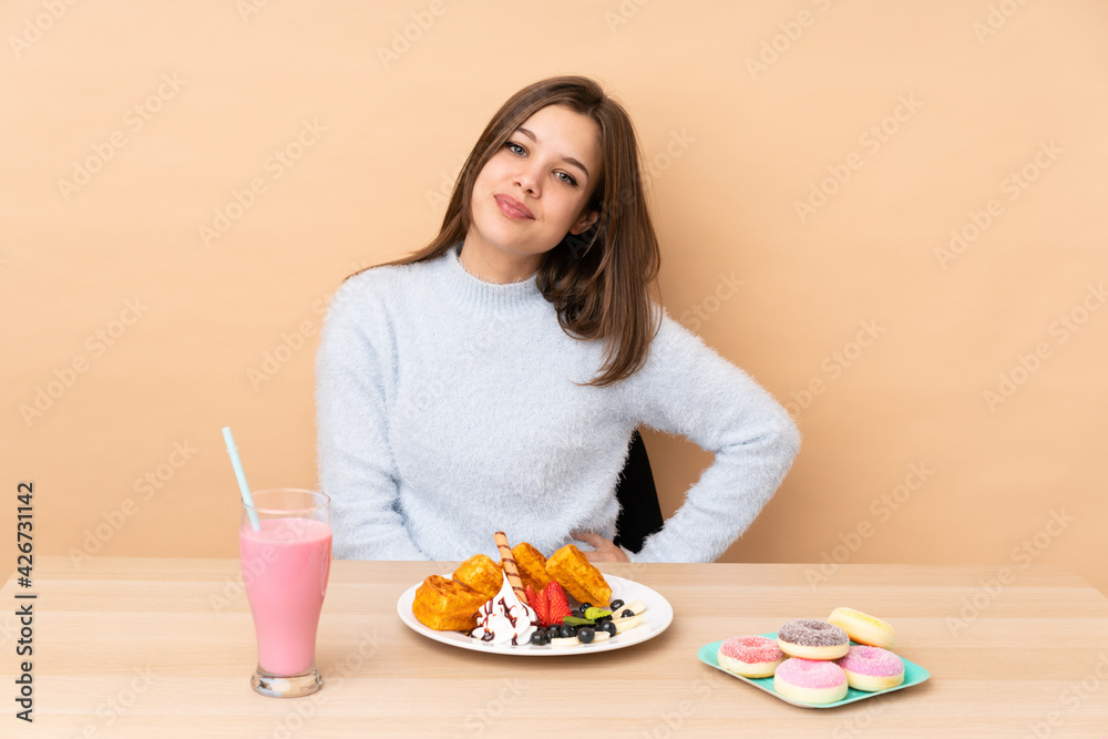 Teenager girl eating waffles isolated on beige background posing with arms at hip and smiling Teenager girl eating waffles isolated on beige background posing with arms at hip and smiling