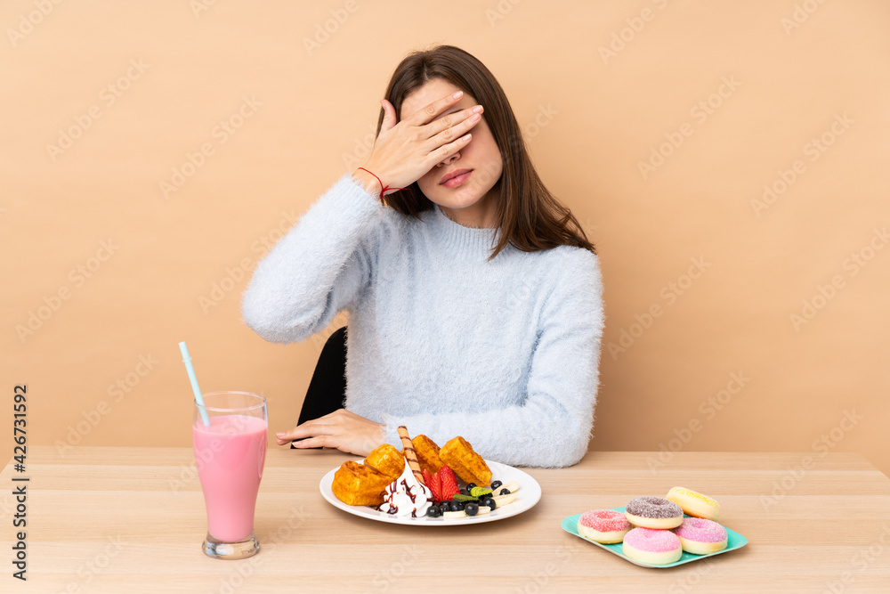 Teenager girl eating waffles isolated on beige background covering eyes by hands. Do not want to see something Teenager girl eating waffles isolated on beige background covering eyes by hands. Do not want to see something