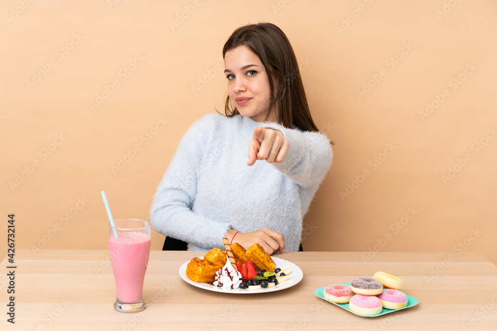 Teenager girl eating waffles isolated on beige background points finger at you with a confident expression Teenager girl eating waffles isolated on beige background points finger at you with a confident expression