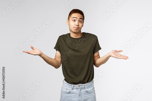 Who knows, I dont aware. Portrait of confused shrugging asian man in greey t-shirt, smirk and apologizing, sorry cant help, standing puzzled and unaware about situation, grey background photo