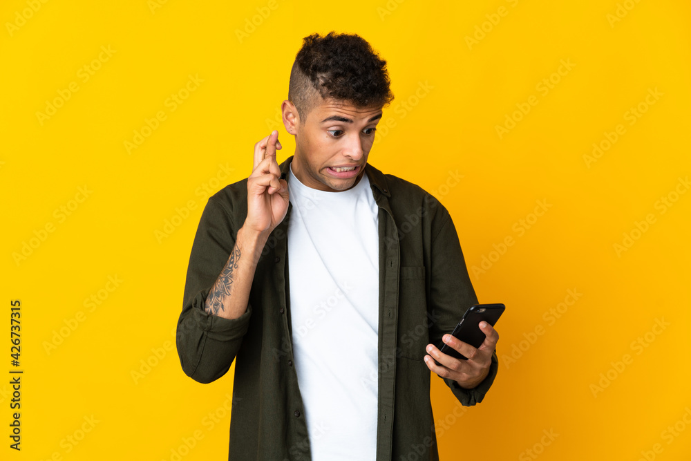 Young brazilian man isolated on yellow background using mobile phone with fingers crossing Young brazilian man isolated on yellow background using mobile phone with fingers crossing