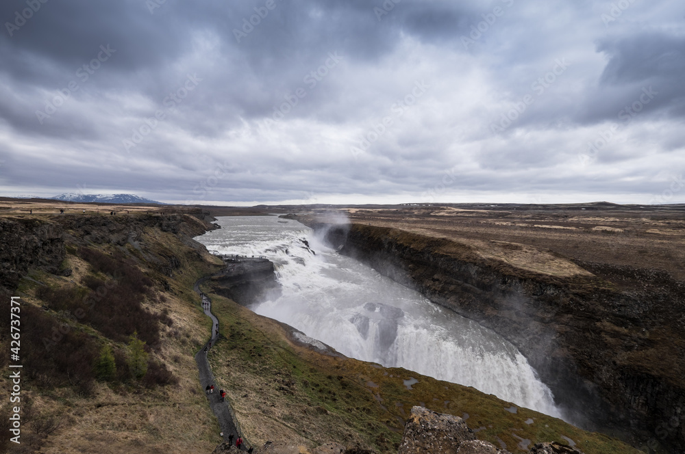 Famous Gulfoss waterfall in the Golden Circle in South Iceland. Unrecognizable tourists walk on a path to the viewpoint. Wide landscape view