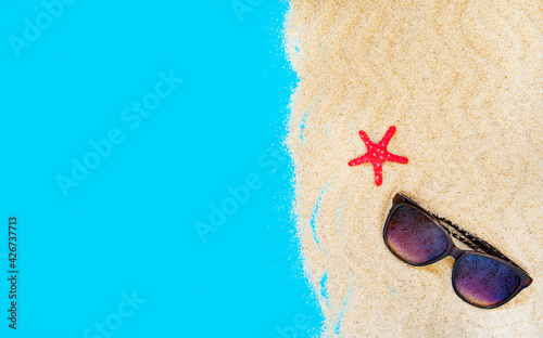 Sand frame with sunglasses and a red starfish on a blue background in the form of the sea or ocean. Flat lay. Top view. Copy space. Vacation, summer and travel concept