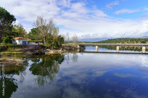 Landscape of a blue lake with trees and green plants  a little house on the shore and a bridge over the water. Atazar Madrid.