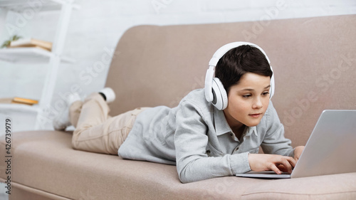 Preteen boy in headphones using laptop on couch.