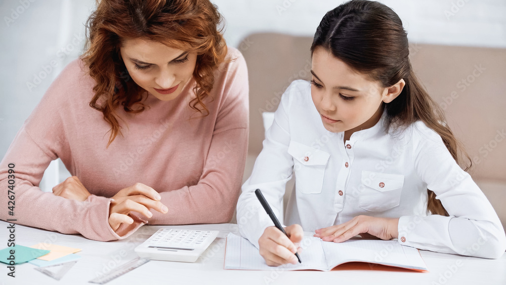 Girl writing on notebook near mother and calculator on table. Girl writing on notebook near mother and calculator on table.