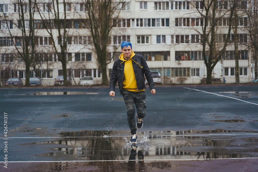 Young man runs through puddle on sports stadium on background of high-rise building. Happy guy with blue hair having fun on city street in springtime.