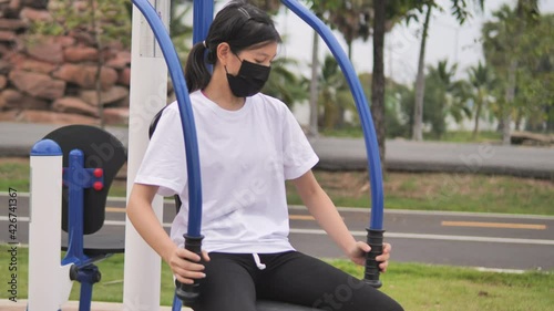 Young woman wearing medical mask is sitting exercise using outdoor exercise equipment in the park. Healthy lifestyle concept.