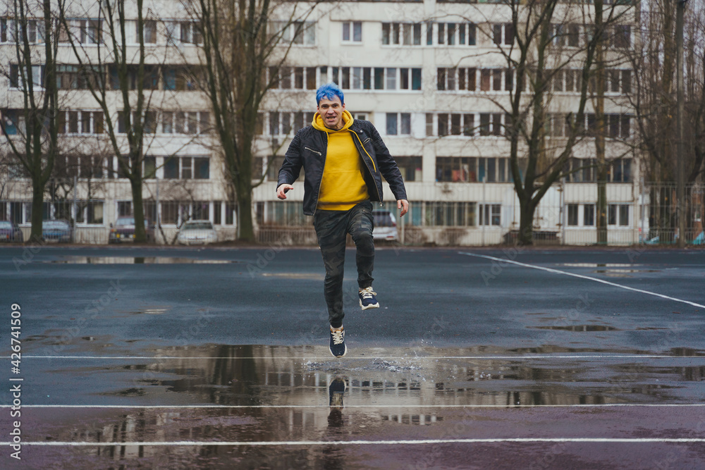 Young man runs through puddle on sports stadium on background of high-rise building. Happy guy with blue hair having fun on city street in springtime.
