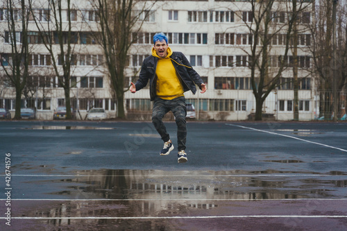 Young man jumps in puddle on sports stadium on background of high-rise building. Happy guy with blue hair having fun on city street in springtime.
