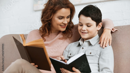 Smiling mother holding book near son with notebook during education at home.