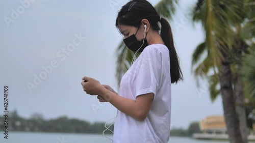 Young woman wearing protective face mask with earphones and are using a smartphone at park. Pandemic and lifestyle concept.