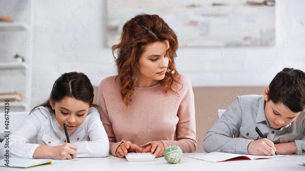 Woman holding calculator near kids writing on notebooks at home. Woman holding calculator near kids writing on notebooks at home.