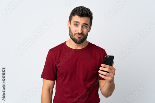 Young handsome man with beard using mobile phone isolated on white background with sad expression